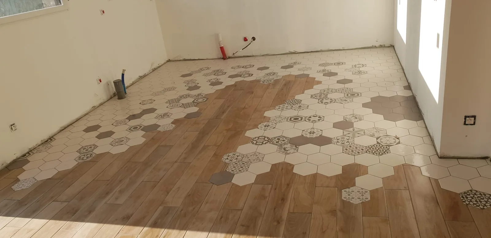 Piece vide vue en grand angle. Le sol associe des lames de carrelage imitation bois et une zone de carreaux hexagonaux clairs a motifs varies, les deux materiaux se melangeant progressivement en degrade.