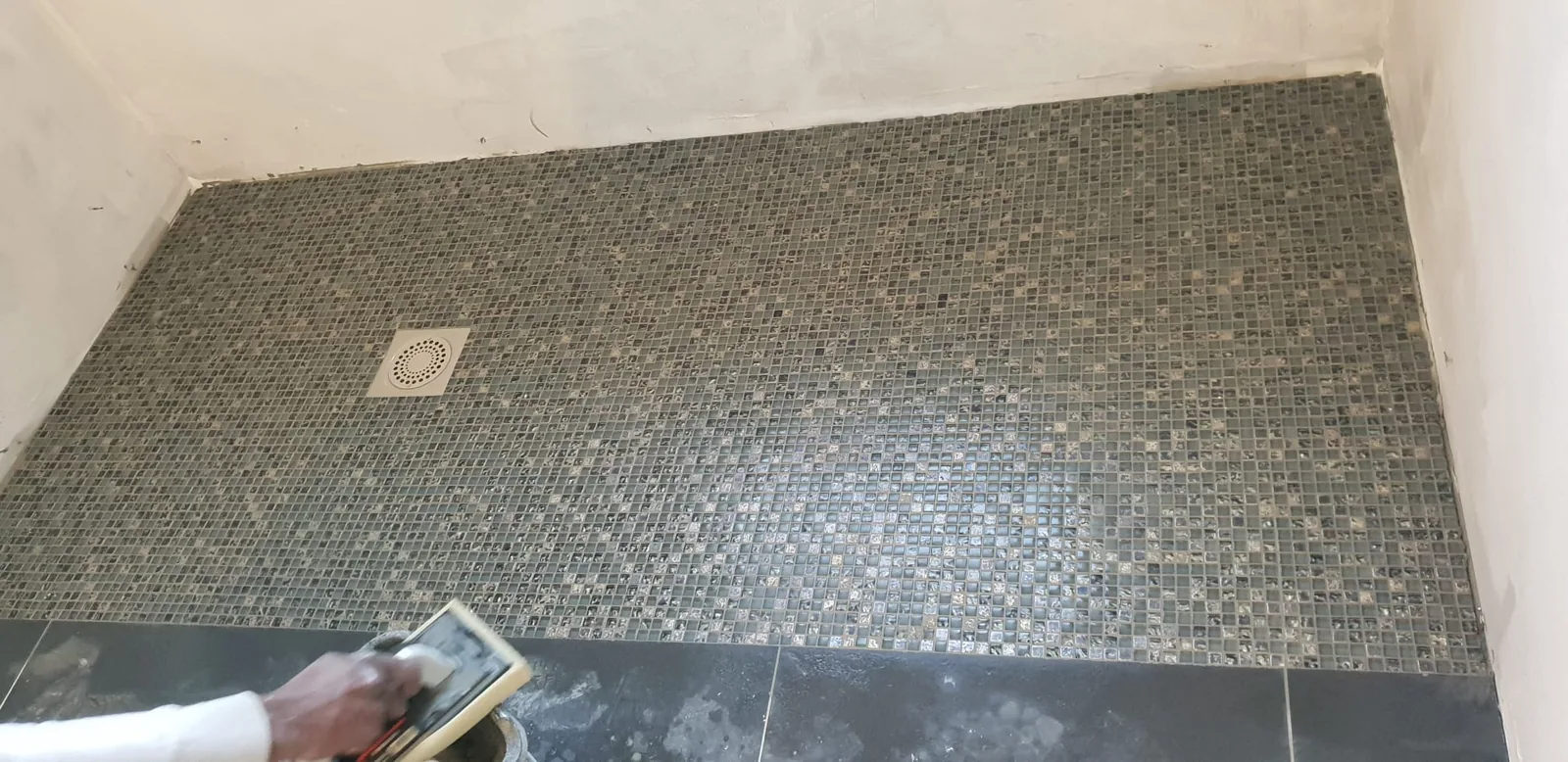 Sol de douche a l'italienne entierement recouvert de mosaique de verre aux tons gris et bruns, avec une bonde carree au centre. En bas de l'image, une main tient une taloche pour appliquer le joint.