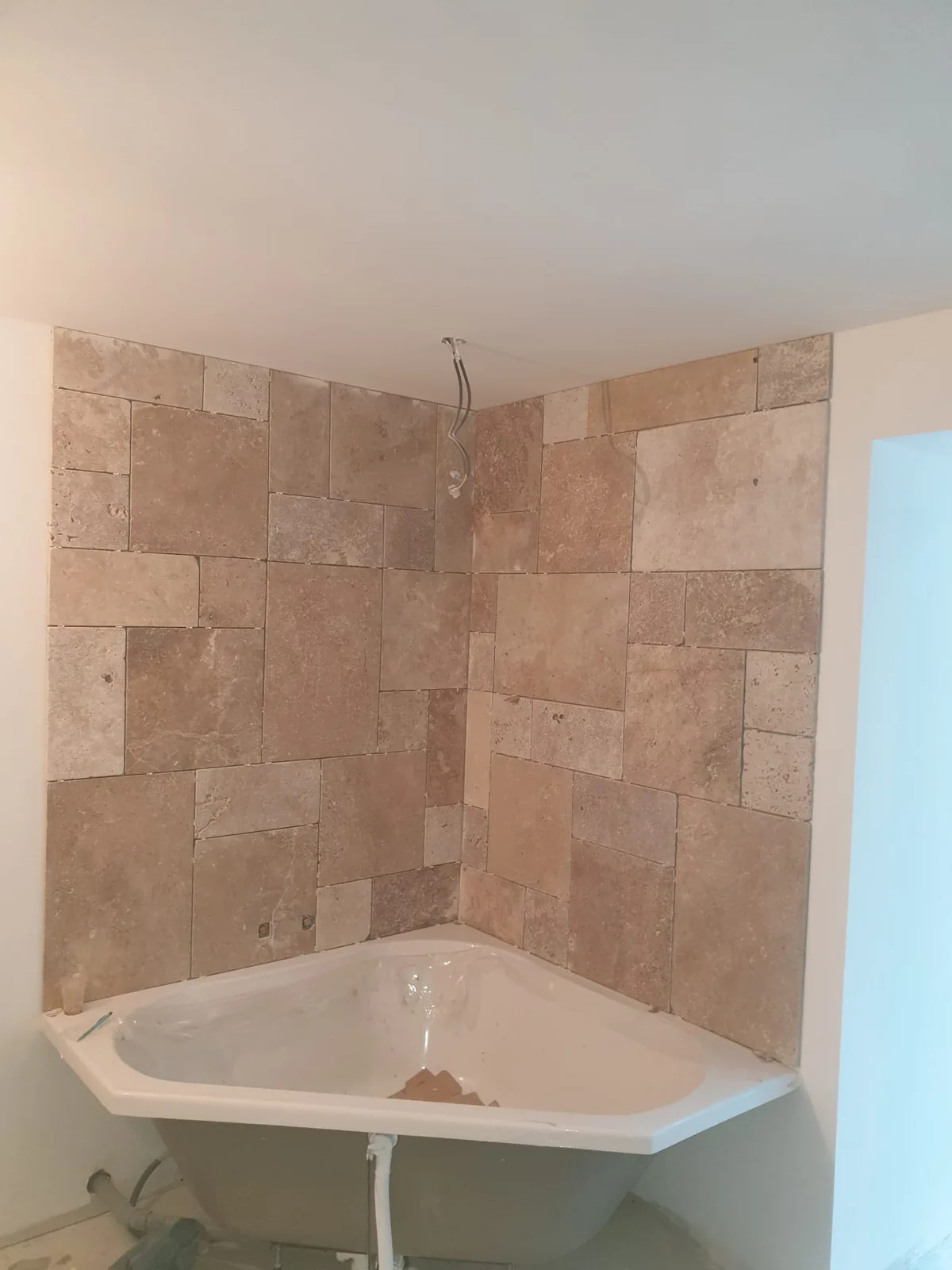 Angle de salle de bains dont les deux murs sont habilles de dalles de travertin beige de formats varies. Une baignoire d'angle blanche est posee au pied du mur, sans habillage, la tuyauterie encore apparente.