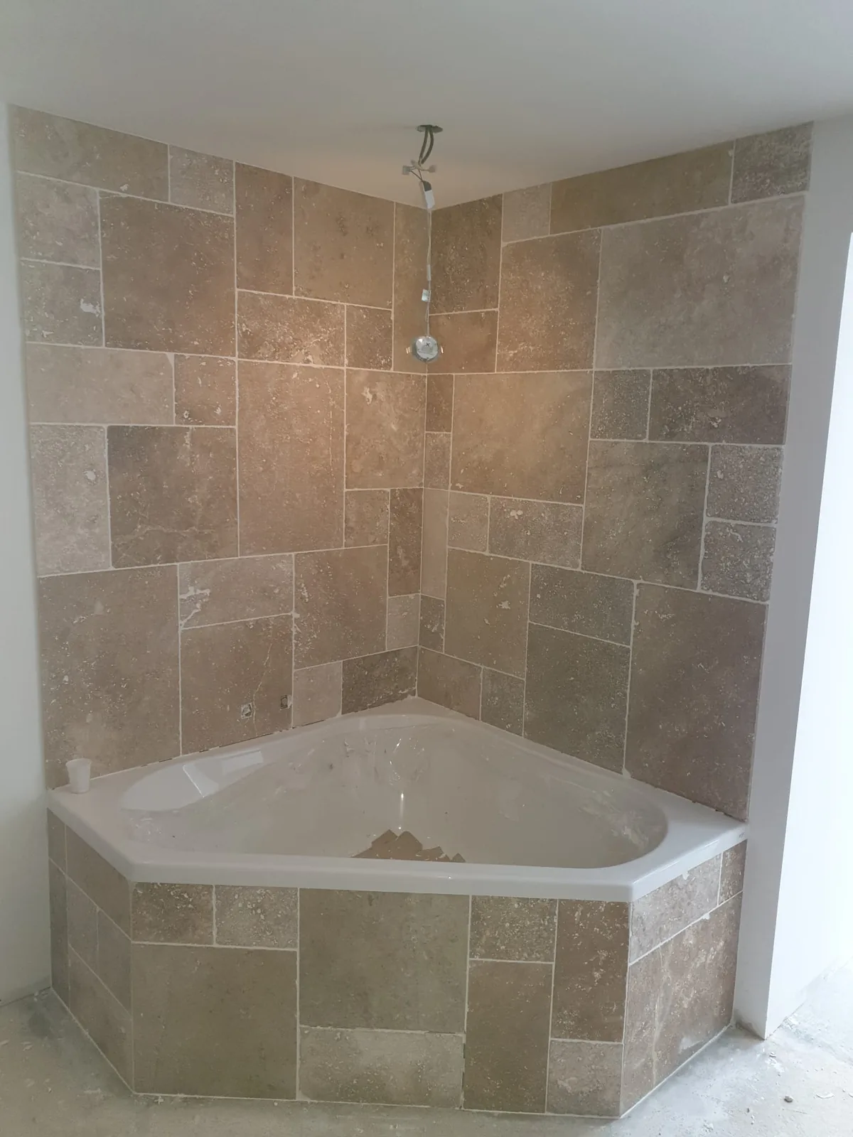 Baignoire d'angle blanche encastree dans un renfoncement entierement habille de travertin beige pose en opus romain, murs et tablier compris. Une douchette est fixee au mur du fond.