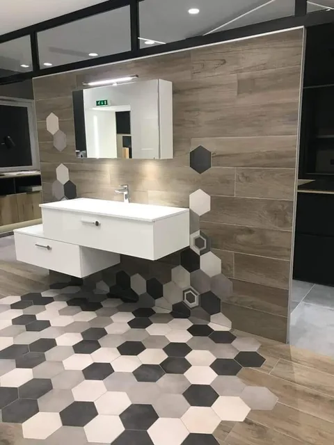 Espace salle de bains avec un meuble vasque blanc suspendu devant un mur habille de lames imitation bois. Des carreaux hexagonaux blancs, gris et noirs habillent le sol et remontent en semis sur le mur.
