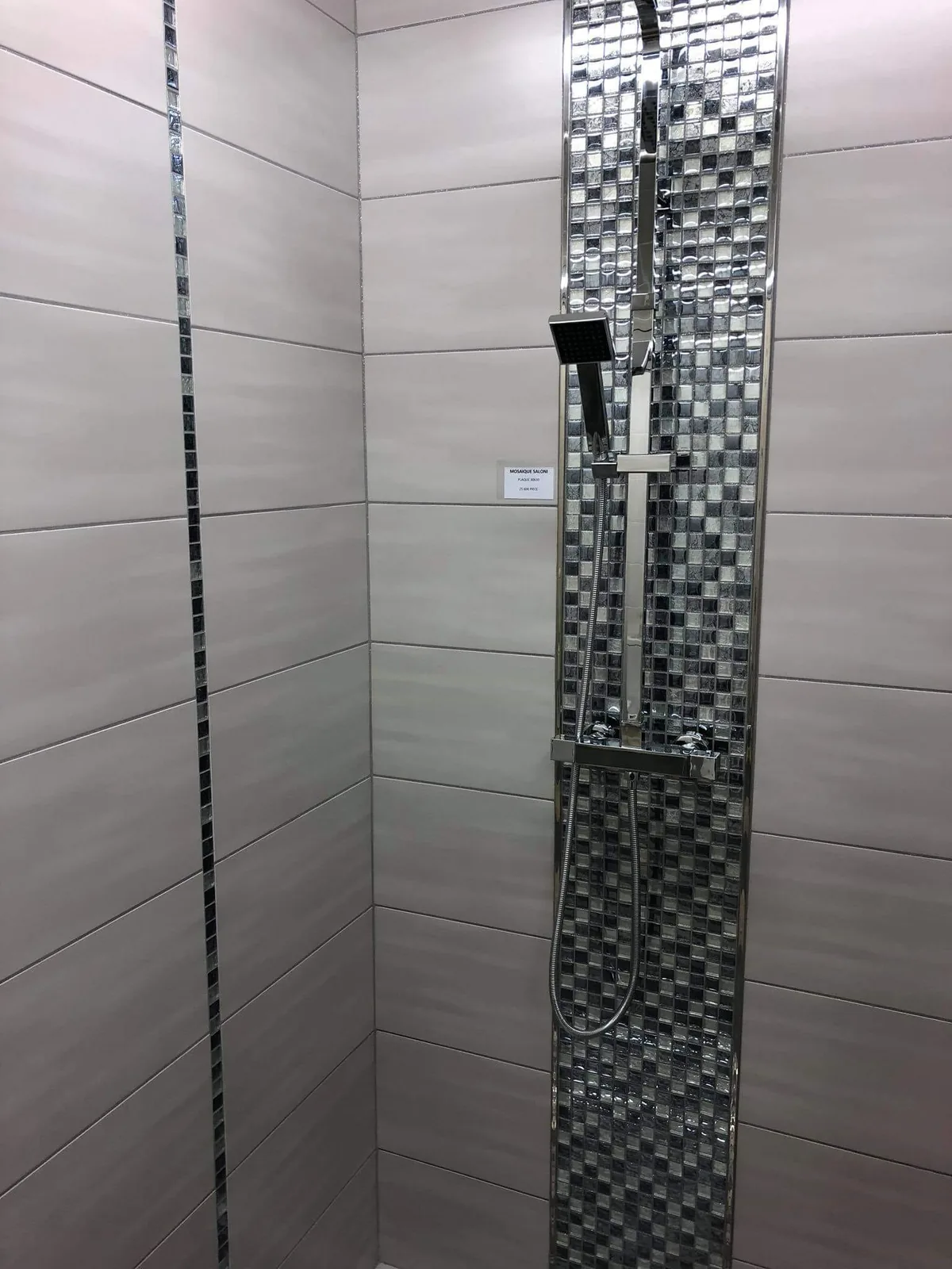 Angle de douche habille de faience grise a lames horizontales, traverse par une fine bande verticale de mosaique de verre. Une large colonne de mosaique de verre argentee accueille la robinetterie et la douchette.