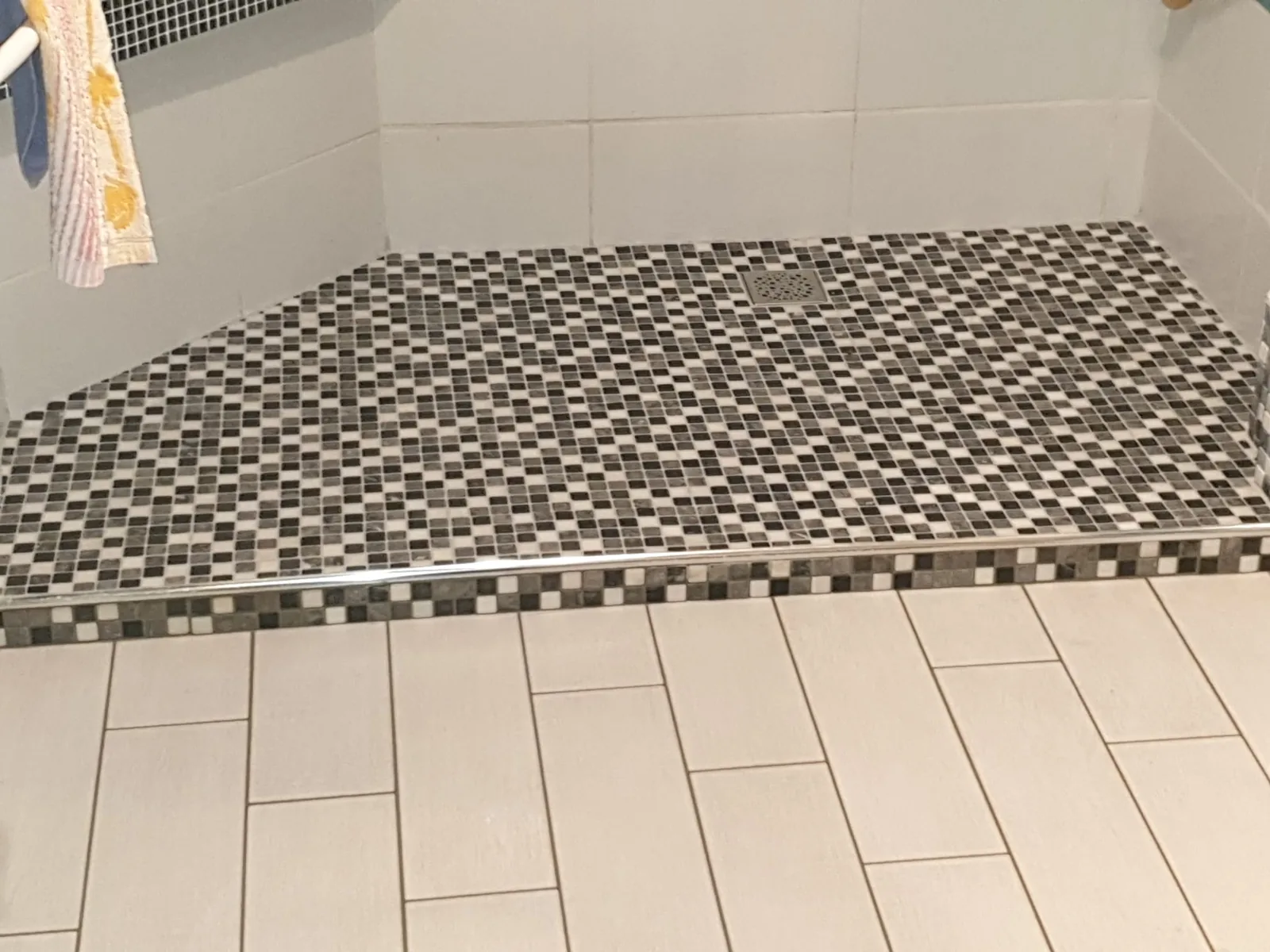 Receveur de douche a l'italienne recouvert de mosaique noire, grise et blanche, ferme par un profil chrome. La meme mosaique forme une frise sur le seuil, devant un sol en carrelage beige clair.