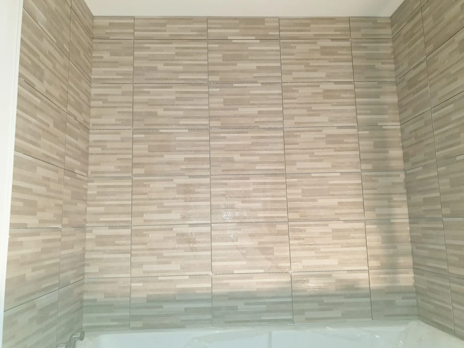 Renfoncement de baignoire dont les trois murs sont habilles d'une faience beige et grise a decor de fines lamelles horizontales. En bas, le rebord d'une baignoire blanche encore protegee par un film plastique.