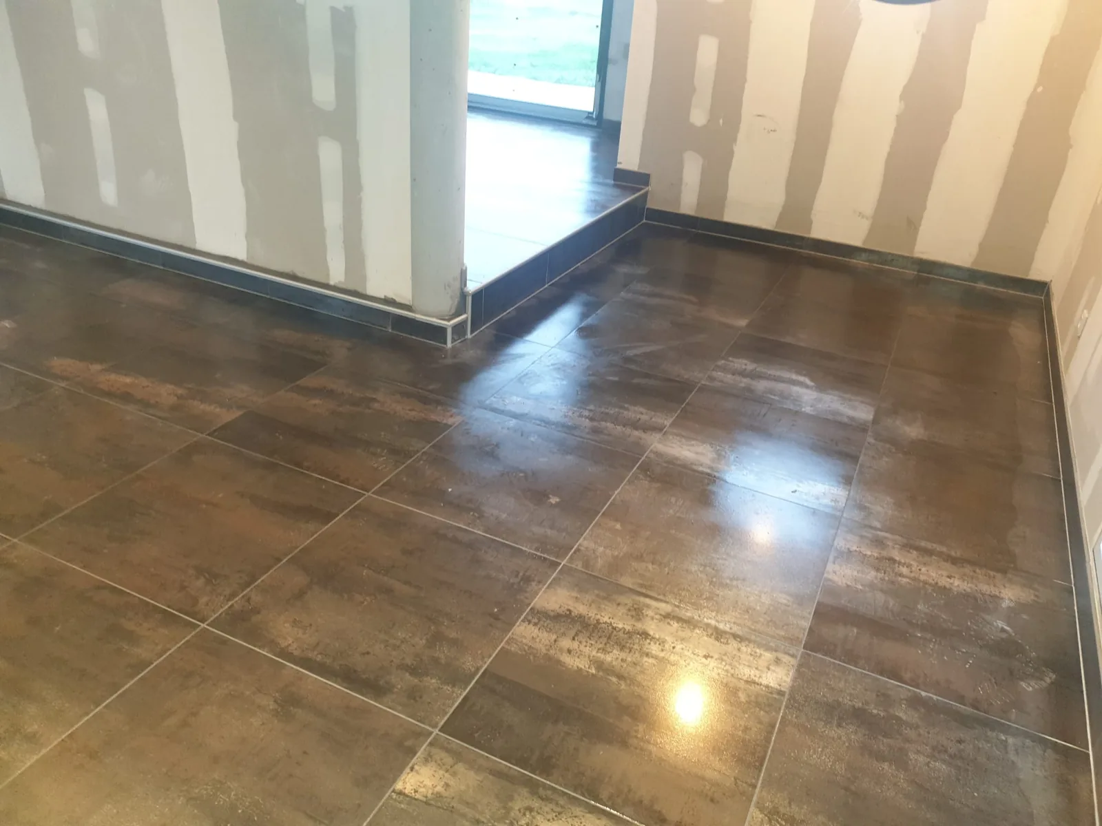 Sol en carreaux bruns brillants de grand format reflechissant la lumiere d'une baie vitree. Une marche habillee de carrelage sombre et un profil metallique bordent le passage vers la piece voisine.
