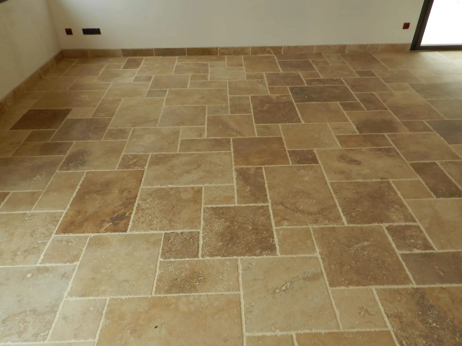Piece vide au sol recouvert de dalles de travertin beige et brun de formats varies, posees en opus romain avec des joints creux clairs. Des plinthes assorties courent au pied des murs blancs.