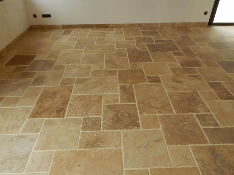 Piece vide au sol recouvert de dalles de travertin beige et brun de formats varies, posees en opus romain avec des joints creux clairs. Des plinthes assorties courent au pied des murs blancs.