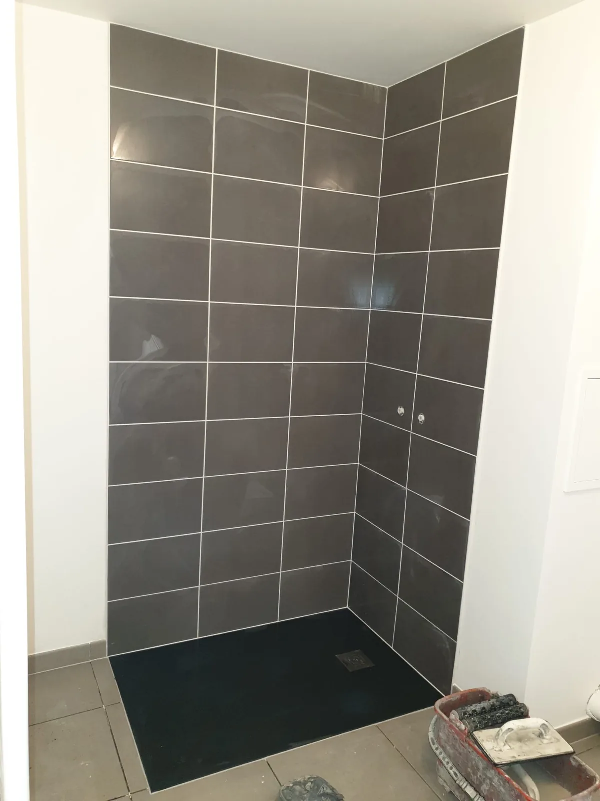 Niche de douche dont les murs sont habilles de faience gris fonce brillante posee droit avec des joints blancs. Le receveur extra-plat noir est installe au sol et deux sorties de robinetterie sont visibles sur le mur de droite.