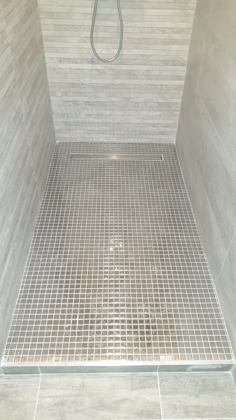 Douche a l'italienne de plain-pied vue en plongee : le sol est entierement recouvert de mosaique grise et beige, ferme au fond par un caniveau lineaire chrome. Les murs sont habilles de carrelage clair effet pierre.