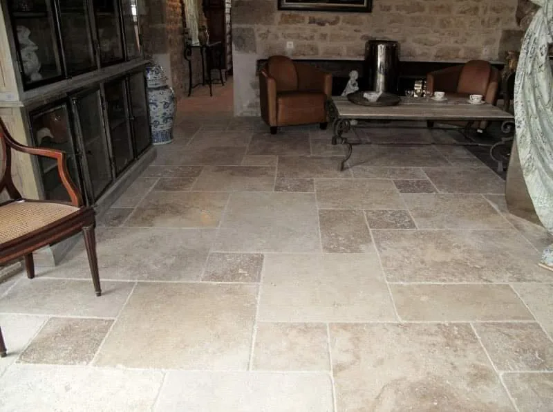 Sejour meuble aux murs en pierre apparente, au sol recouvert de dalles de travertin clair de formats varies posees en opus romain. Au fond, deux fauteuils et une table basse ancienne ; a gauche, une vitrine vitree.