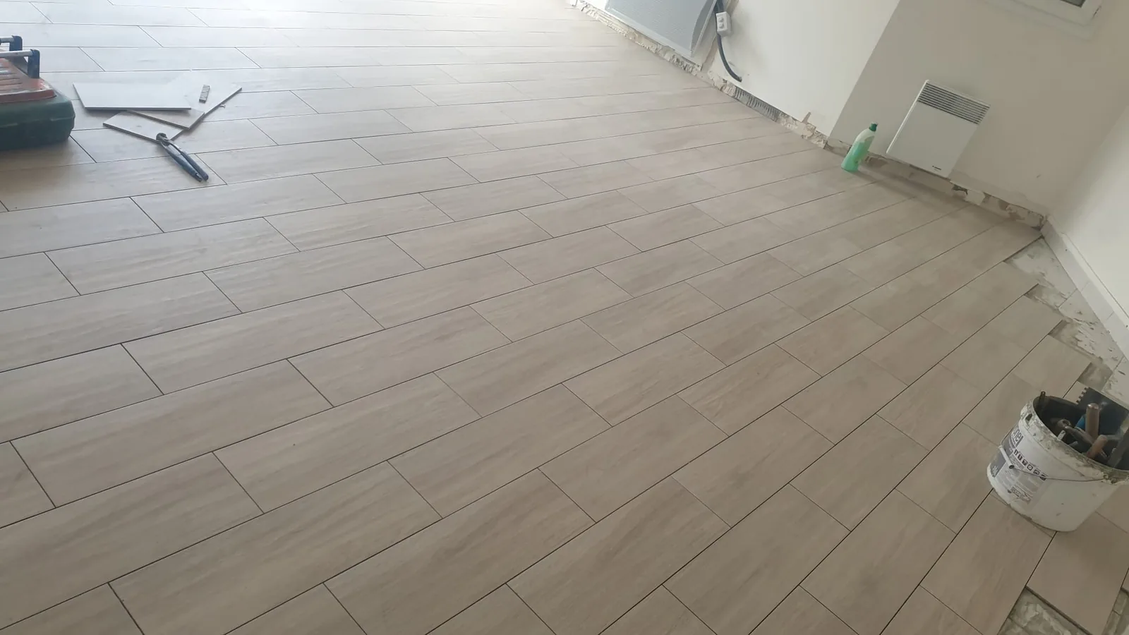 Piece au sol recouvert de lames de carrelage imitation bois beige clair posees a coupe decalee. Une caisse a outils et une regle sont posees en haut a gauche, un seau d'outils en bas a droite.
