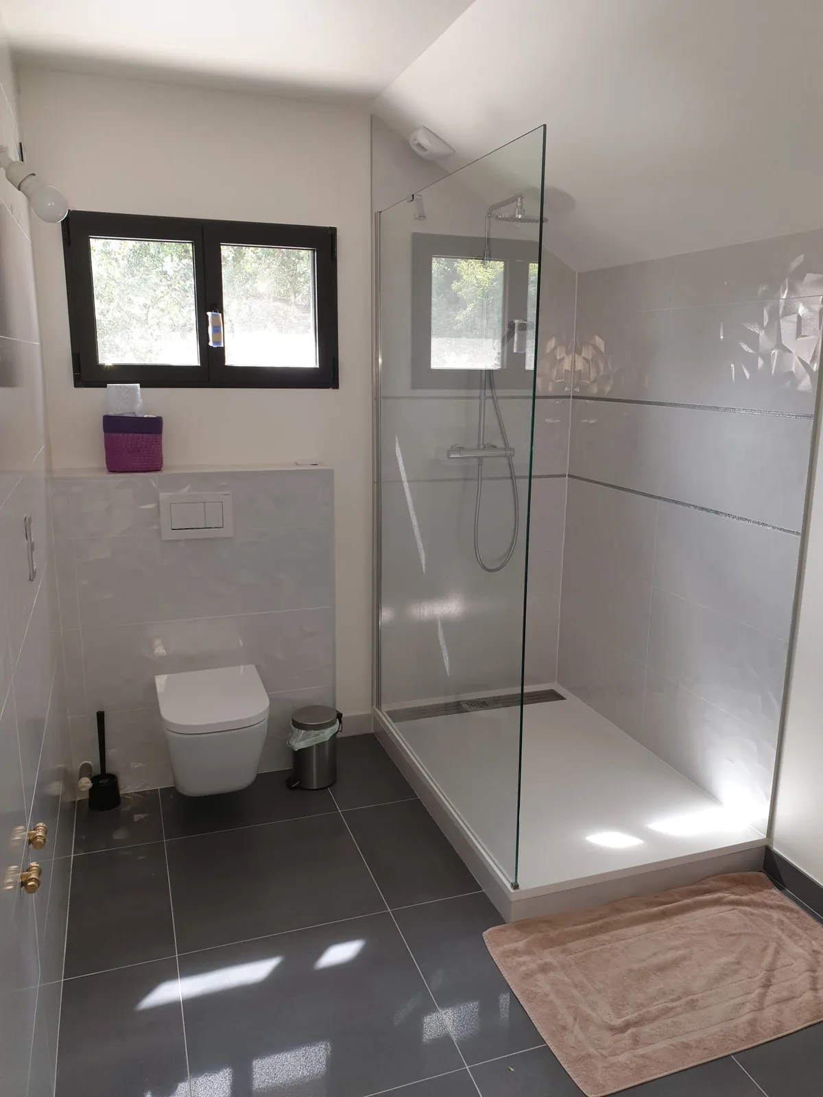 Salle d'eau carrelée terminée avec douche à l'italienne, paroi en verre, WC suspendu, faïence blanche en relief et sol en carrelage gris grand format