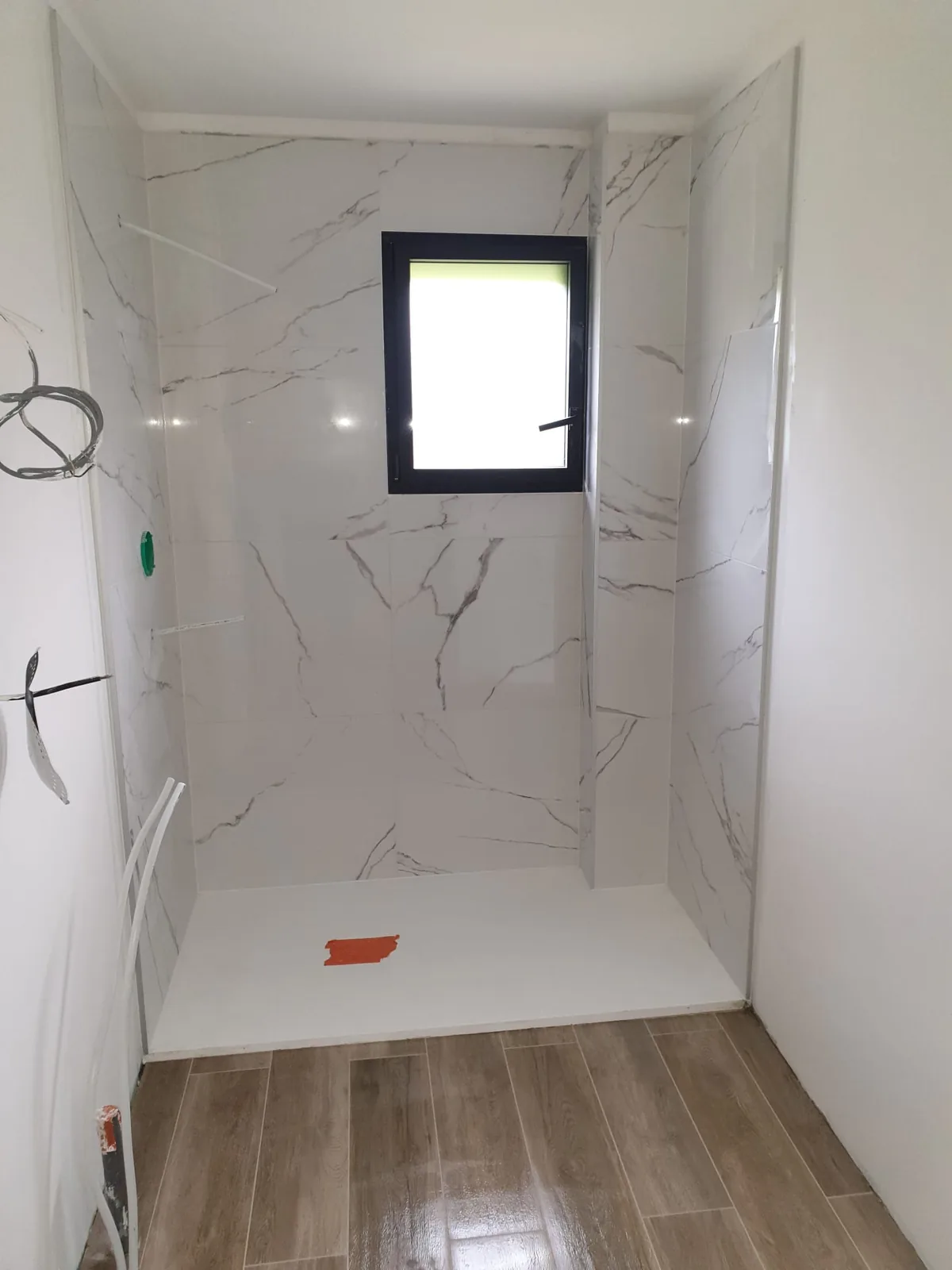 Douche à l'italienne carrelée en grand format blanc effet marbre, avec petite fenêtre à cadre noir et sol en carrelage imitation bois