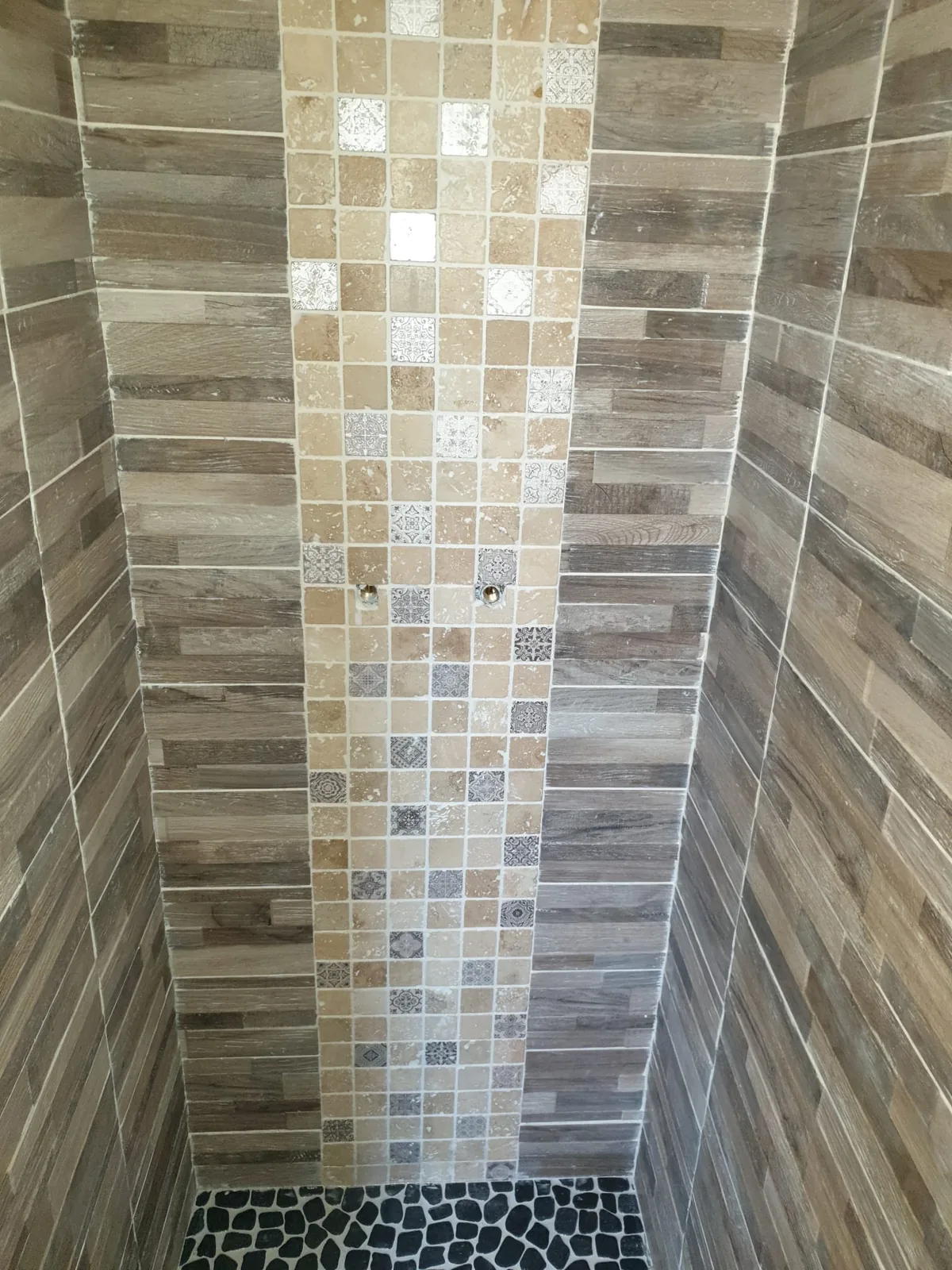 Intérieur de douche à l'italienne avec murs en carrelage imitation bois, bande décorative verticale en mosaïque de pierre à inserts à motifs et sol en galets noirs