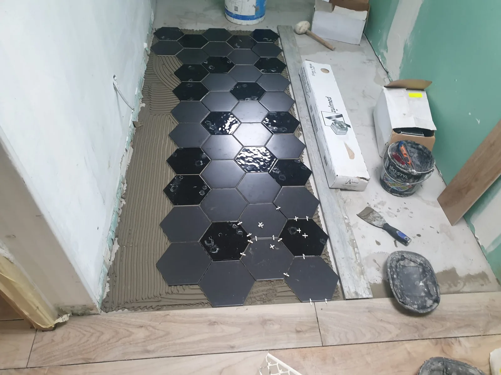 Chantier de pose de carrelage hexagonal noir sur colle peignée, avec croisillons, seaux et outils autour