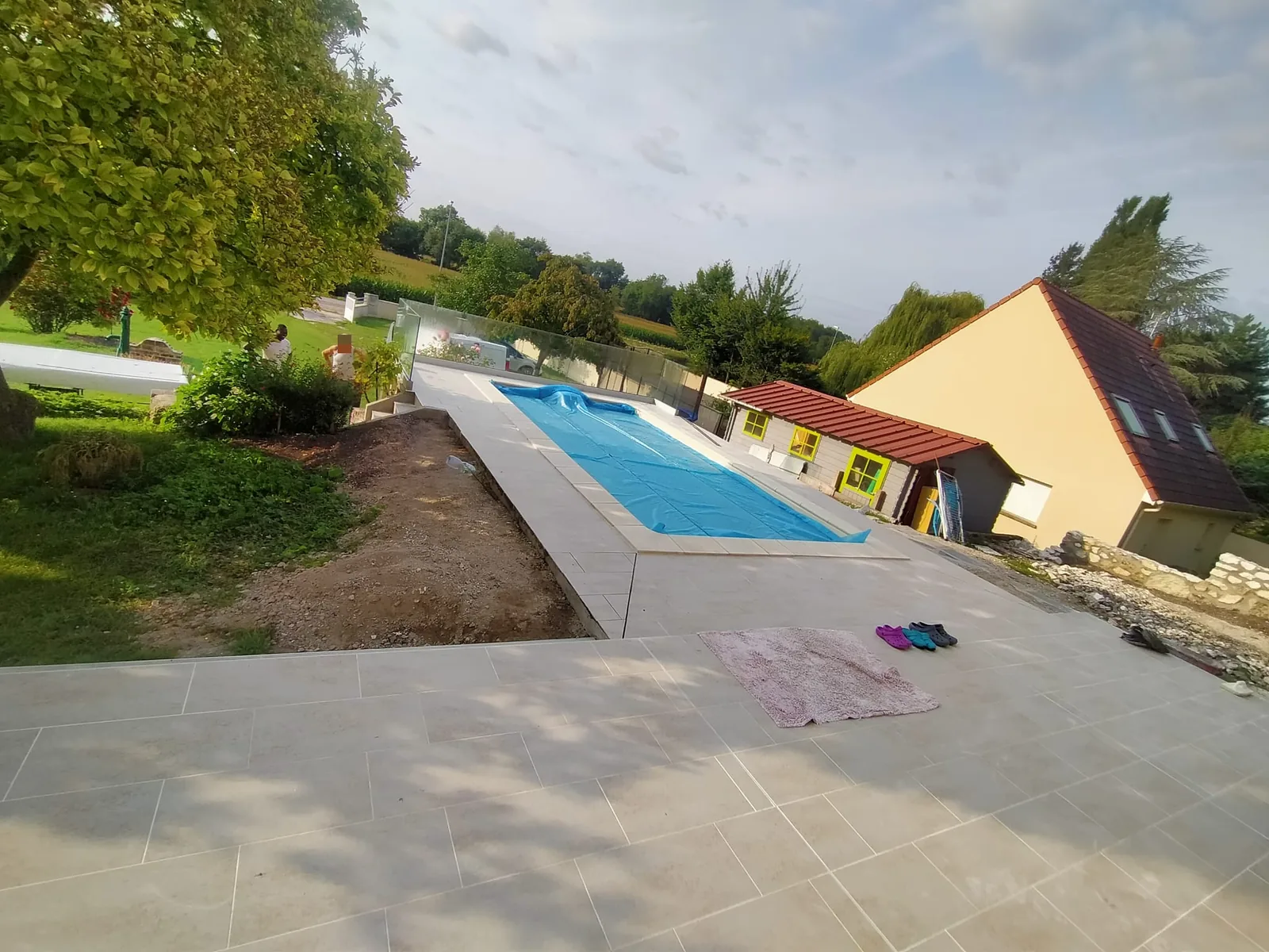 Piscine enterrée avec couverture bleue, entourée d'une plage carrelée en grandes dalles claires, dans un jardin arboré