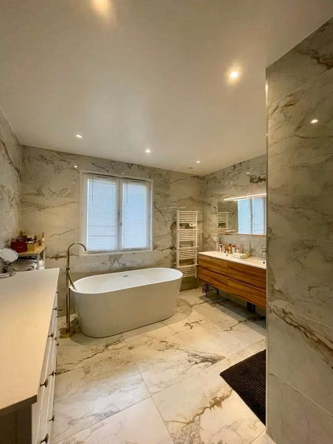 Salle de bains terminee entierement revetue de carrelage grand format effet marbre veine beige et gris, avec baignoire ilot blanche, robinetterie sur pied doree, meuble vasque suspendu en bois et seche-serviettes