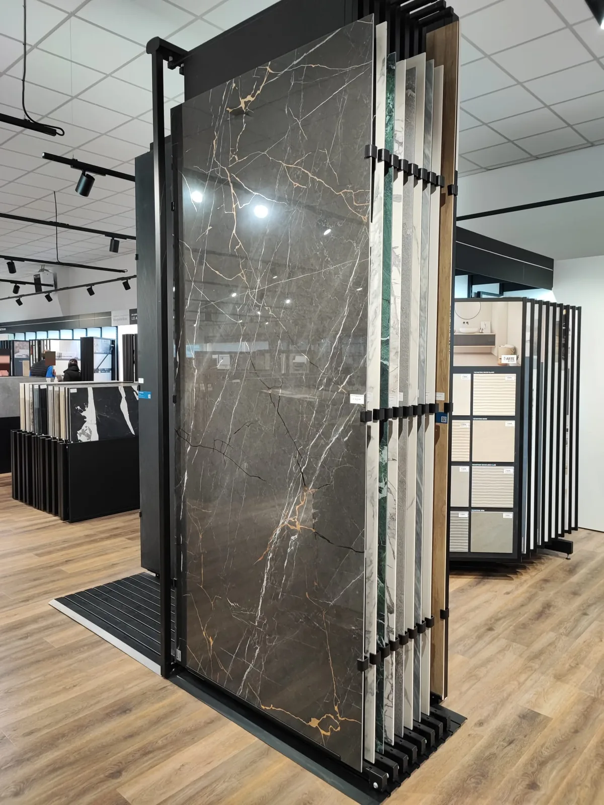 Presentoir vertical a volets coulissants presentant de grandes dalles ceramiques effet marbre, dont une dalle sombre veinee de blanc et d'ocre au premier plan, dans une salle d'exposition eclairee par des spots sur rail