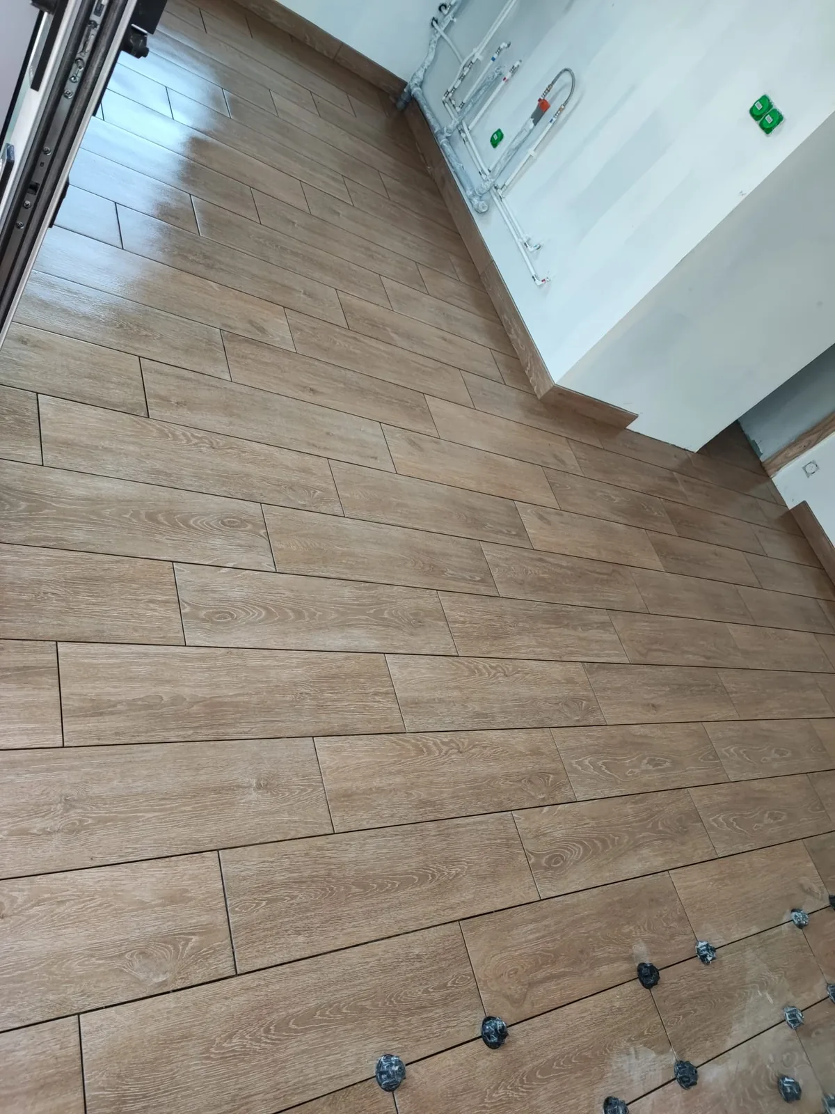 Vue en plongee d'un sol en carrelage imitation bois pose a joints decales, avec cales autonivelantes noires encore en place au premier plan, dans une piece en chantier