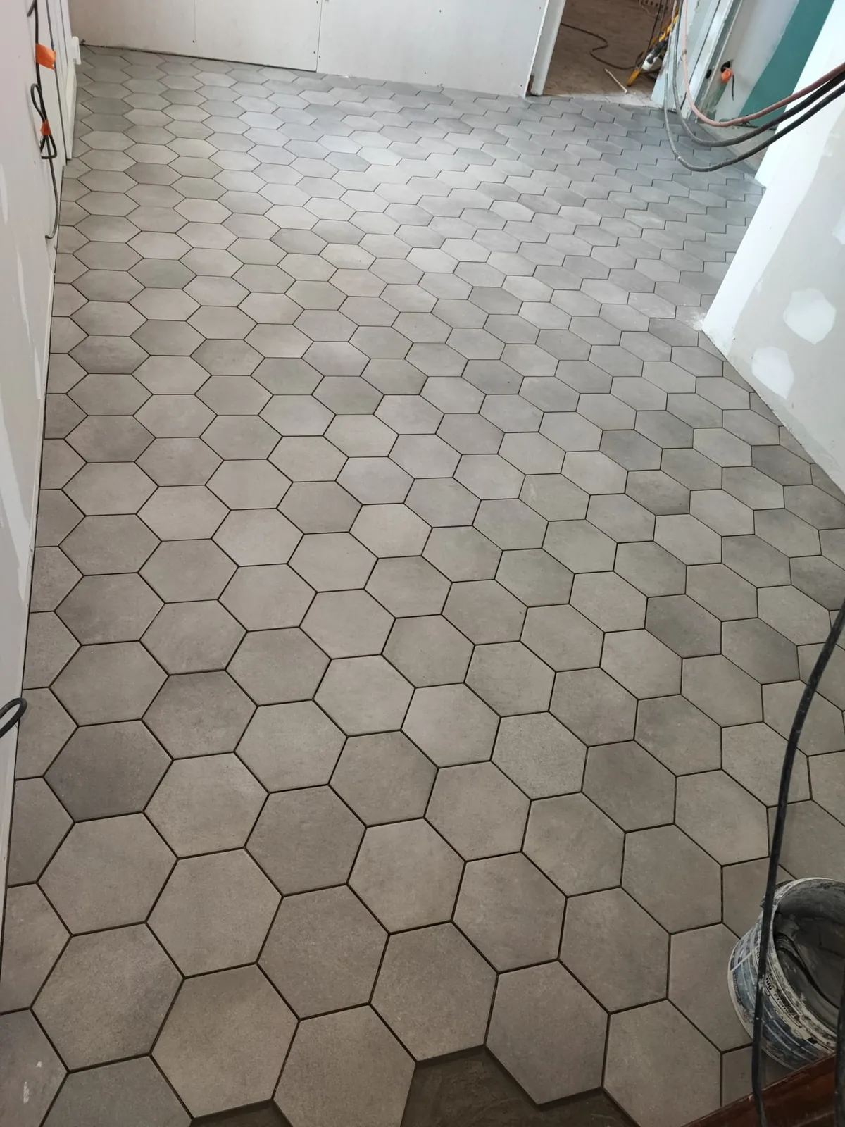 Sol entierement recouvert de carreaux hexagonaux gris clair effet beton, pose en nid d'abeille avec joints reguliers, dans une piece dont les murs en plaques de platre ne sont pas encore finis