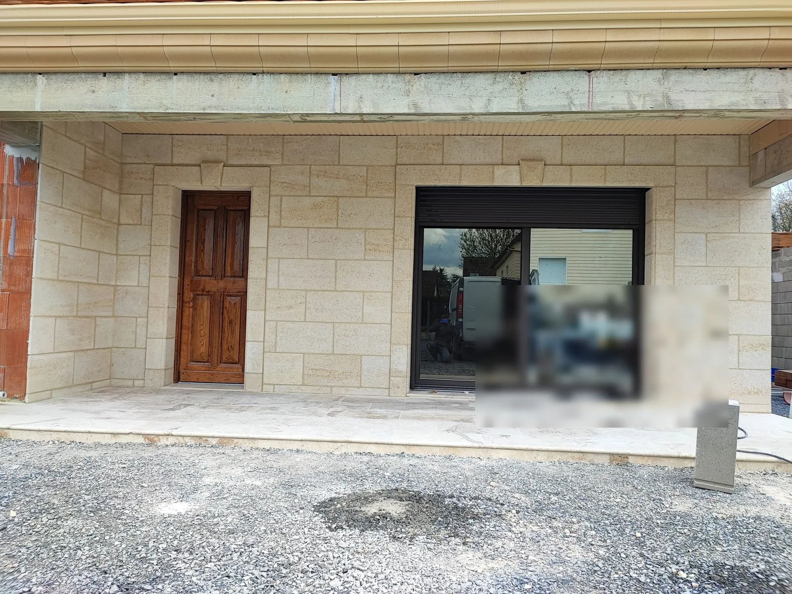 Facade de plain-pied en cours de finition, habillee de plaques de parement effet pierre de taille beige avec encadrements et clef de voute, porte en bois vernis et grande baie coulissante a menuiserie sombre, terrasse en dalles claires devant