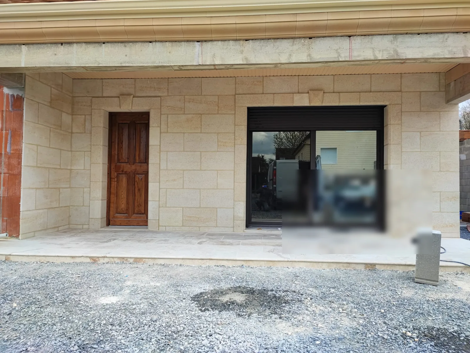 Facade de maison de plain-pied habillee de parement effet pierre de taille beige avec encadrements et clef de voute, porte en bois vernis et baie coulissante sombre, dalles de terrasse claires au sol et gravier au premier plan