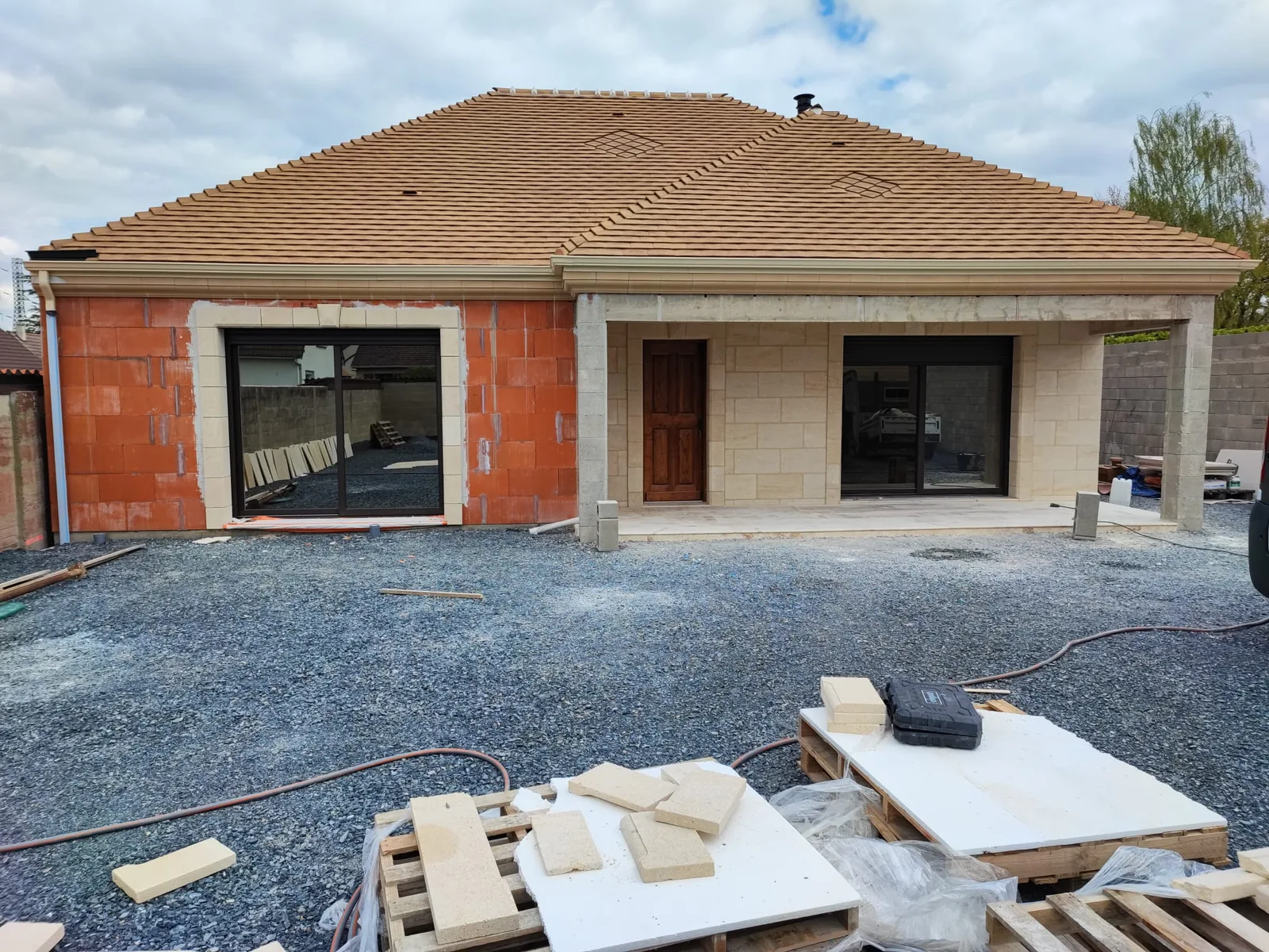 Maison de plain-pied a toiture en tuiles, dont la facade est partiellement habillee de parement effet pierre de taille : une partie du mur est encore en briques rouges, palettes de plaques de parement et outils au premier plan sur le gravier