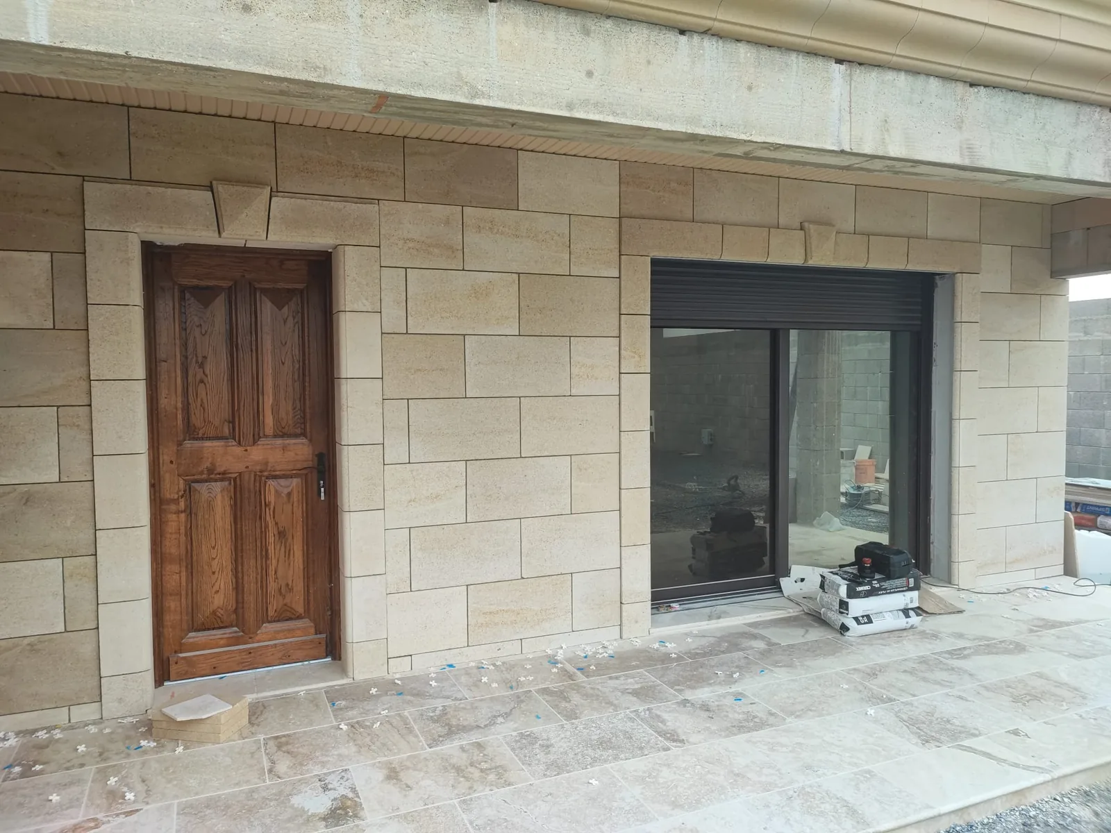 Facade habillee de plaques de parement effet pierre de taille beige avec encadrements de porte et de baie, porte en bois vernis, et terrasse en dalles beiges veinees dont les croisillons de pose sont encore en place au sol