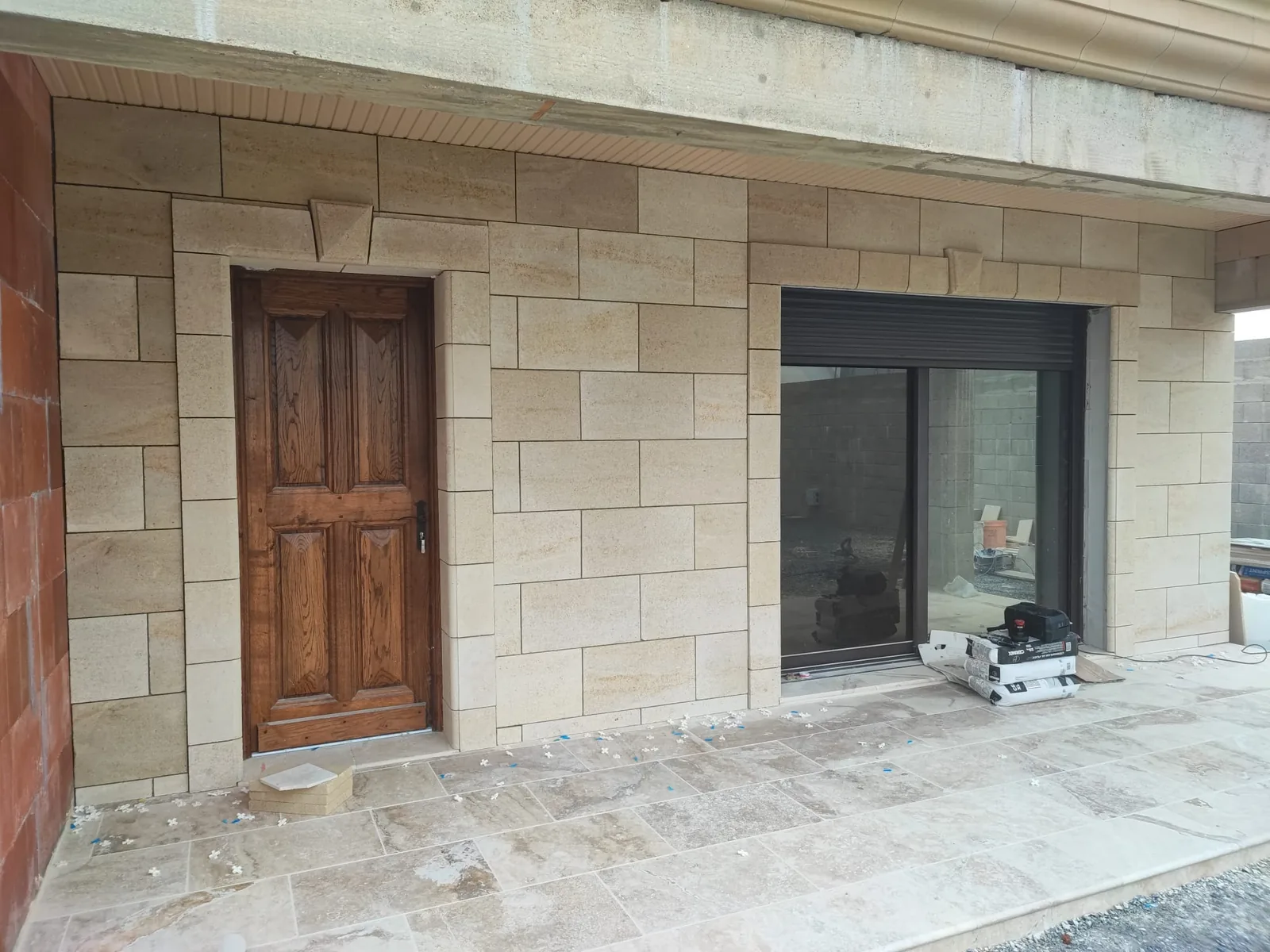 Vue de trois quarts d'une facade habillee de parement effet pierre de taille beige avec encadrements et clefs de voute, porte en bois vernis, baie coulissante sombre et terrasse en dalles beiges veinees avec croisillons de pose