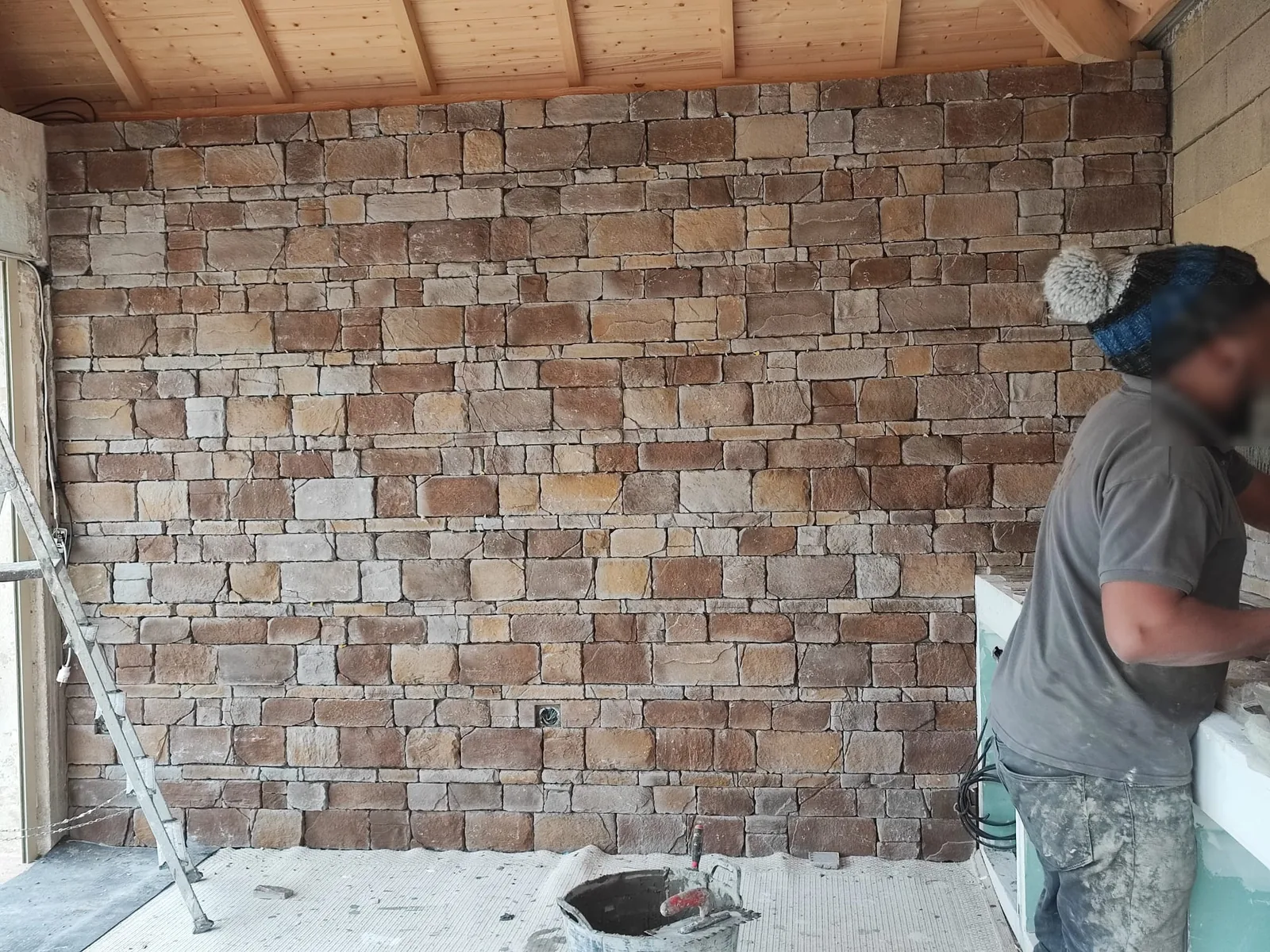 Grand mur habille de parement pierre aux tons bruns, ocres et gris en assises irregulieres, sous une charpente en bois clair apparente, dans un espace couvert en chantier avec escabeau et seau de colle au sol
