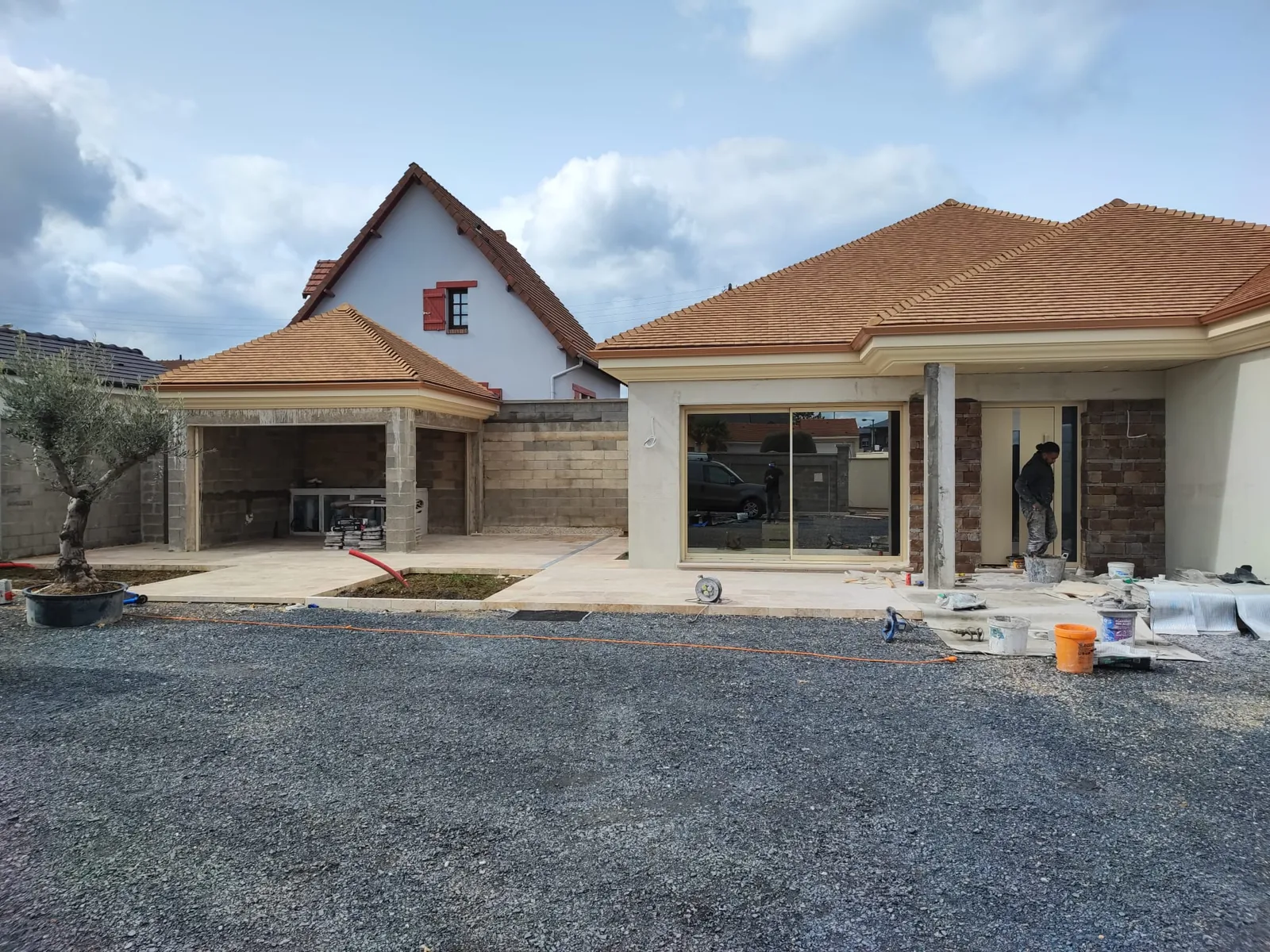 Vue large d'une maison en chantier avec terrasse en dalles beiges, grande baie coulissante, murs habilles de parement pierre fonce, espace couvert a gauche encore en parpaings et olivier en pot, sur une cour en gravier