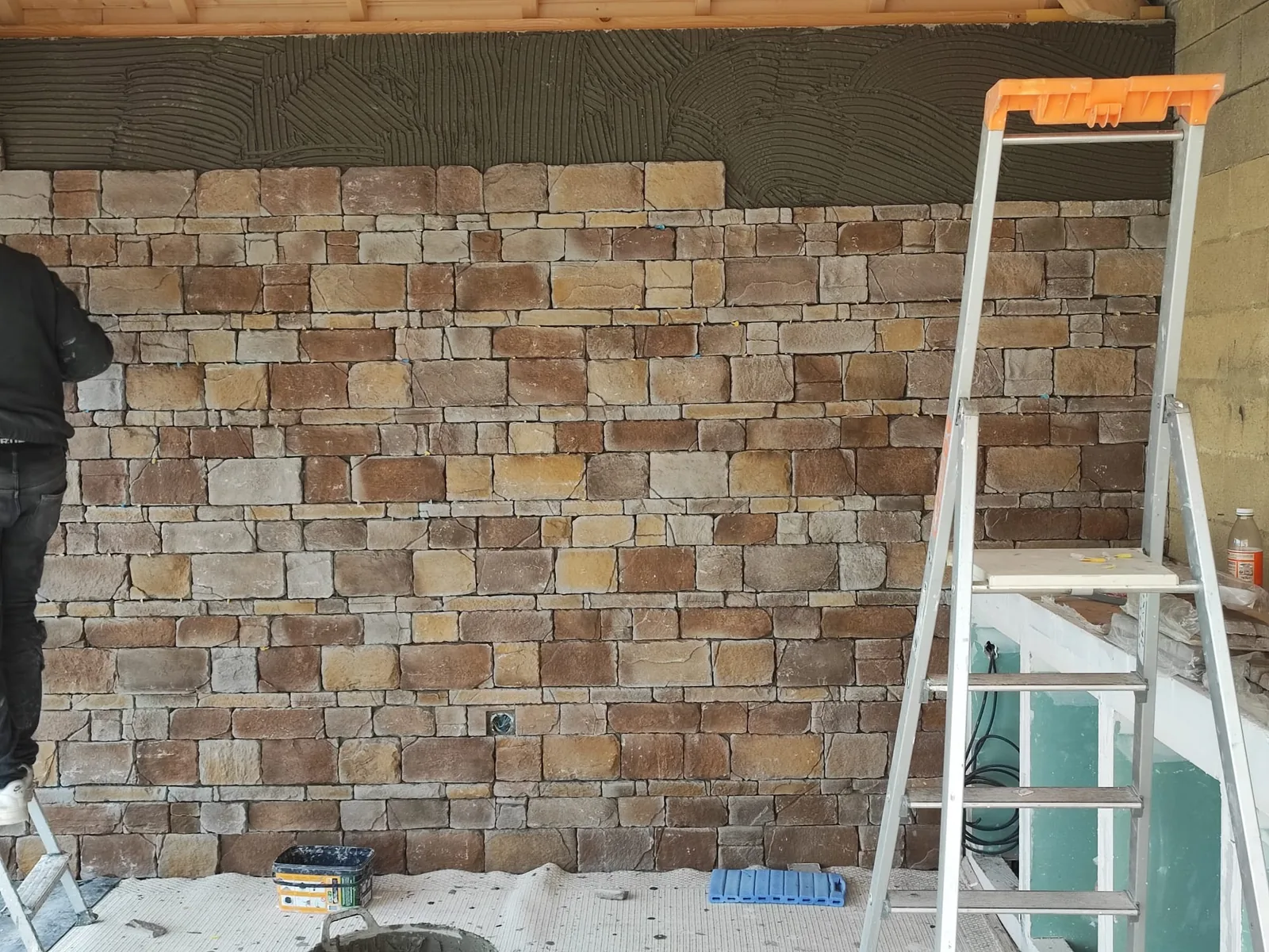 Mur en cours d'habillage de parement pierre aux tons bruns et ocres : la partie haute laisse voir la colle peignee verte pretes a recevoir les dernieres pierres, avec un escabeau a droite et un seau au sol