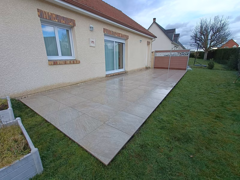 Terrasse rectangulaire en grands carreaux beiges et gris aspect pierre, encore mouillee par la pluie, longeant la facade en crepi clair d'une maison de plain-pied et bordee de pelouse