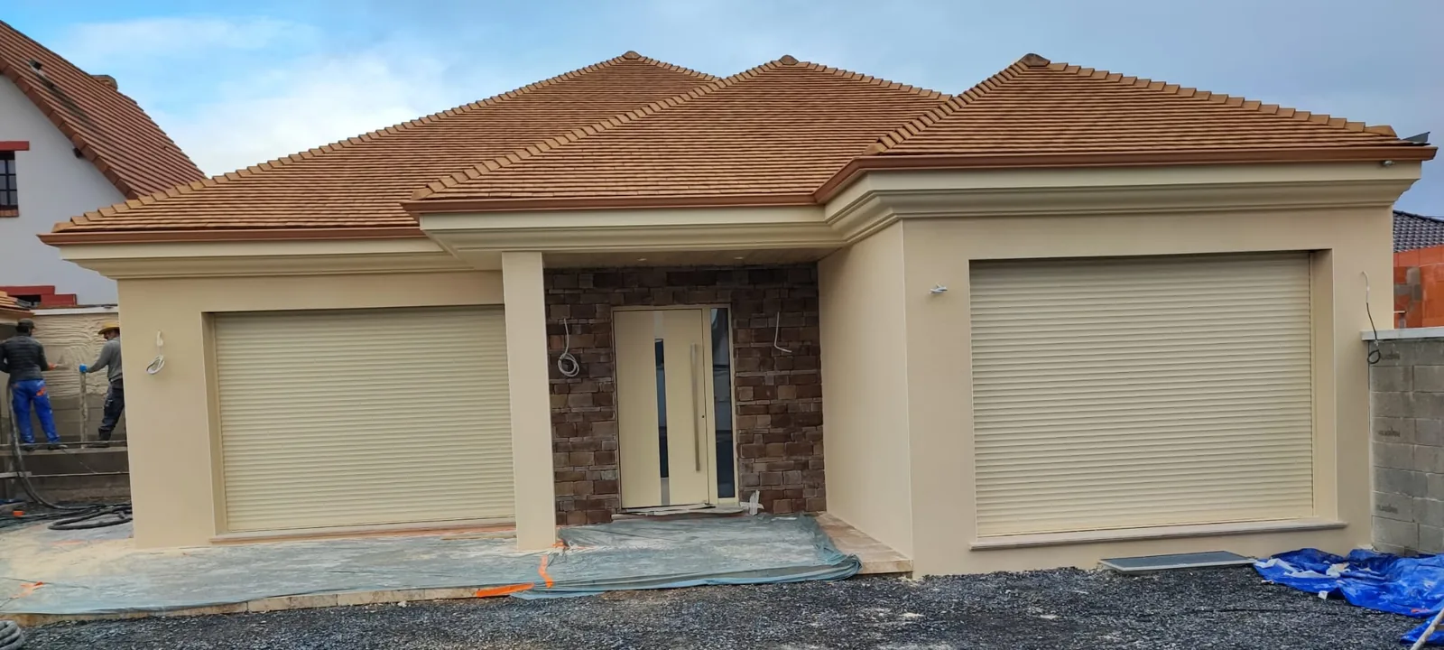 Facade panoramique d'une maison de plain-pied a enduit beige et toiture en tuiles, avec deux grands volets roulants clairs et un renfoncement d'entree habille de parement pierre brun fonce encadrant une porte claire