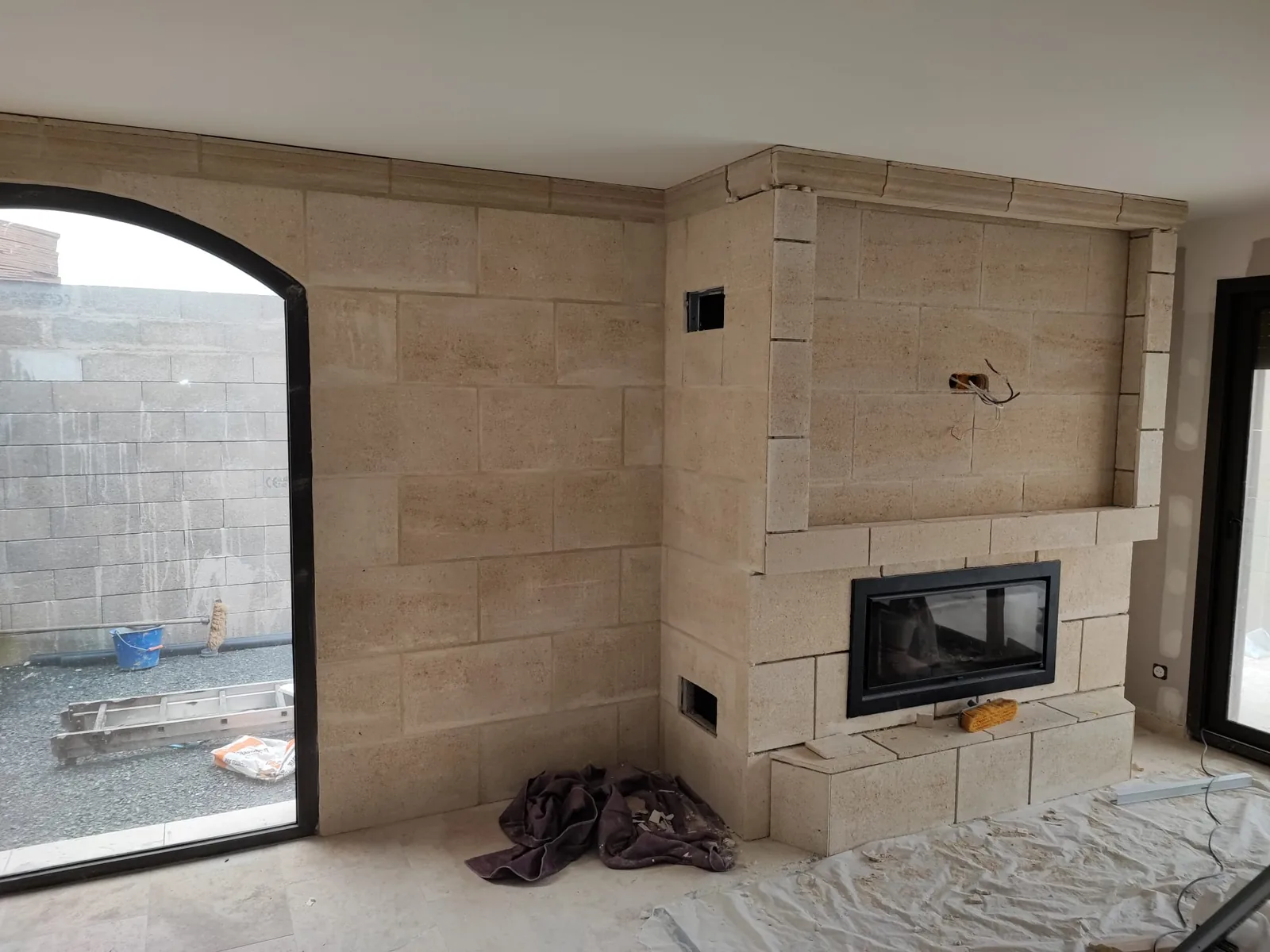 Angle d'une piece en chantier dont le mur et le manteau de cheminee sont habilles de parement effet pierre de taille beige, avec un insert de cheminee vitre encastre, une baie cintree a gauche donnant sur le chantier et des baches de protection au sol