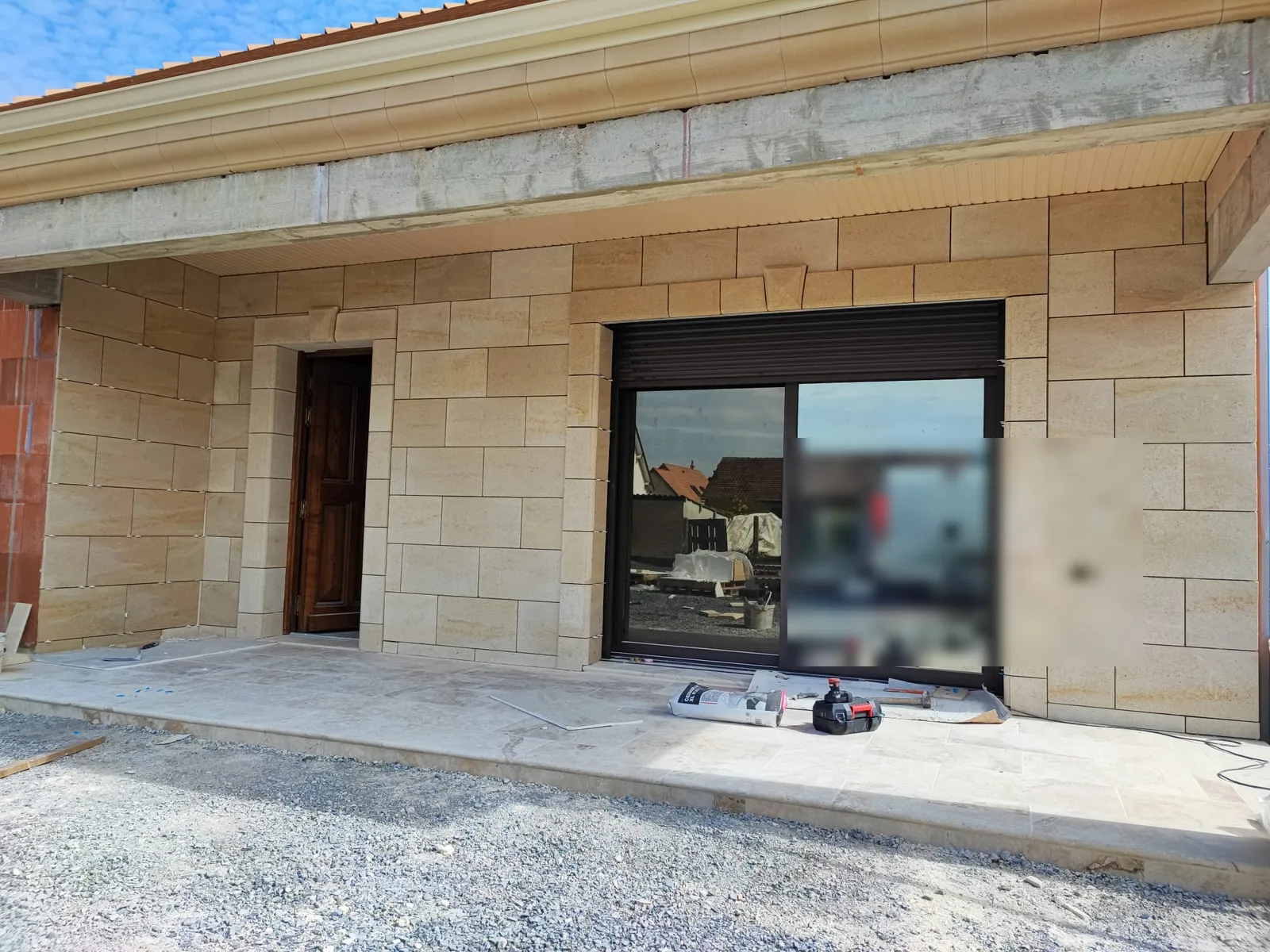 Facade habillee de parement effet pierre de taille beige avec encadrements de porte et de baie, porte en bois vernis entrouverte, baie coulissante sombre et terrasse en dalles beiges, sous un ciel bleu