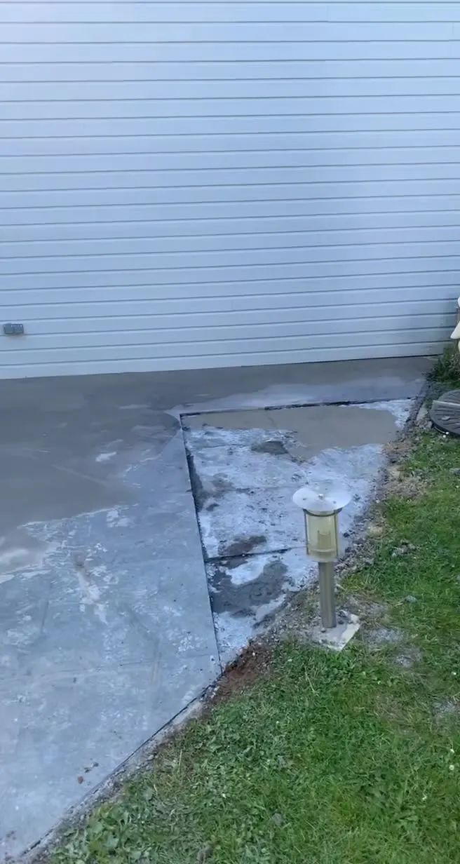 Dalle de béton gris en bordure d'une maison à bardage clair, pelouse et petite borne d'éclairage sur le côté