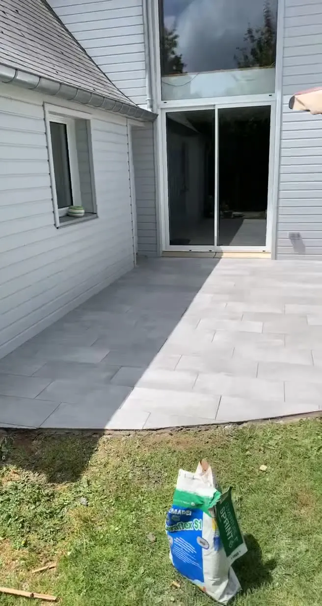 Terrasse extérieure en dalles grises posées à joints décalés devant une baie vitrée coulissante, pelouse au premier plan