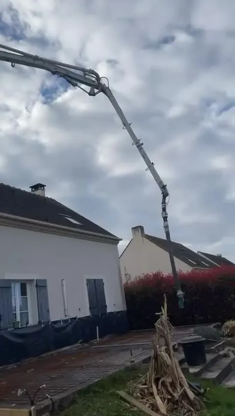 Bras articule d'une pompe a beton deploye au-dessus d'un terrain en terre, devant une maison individuelle en chantier